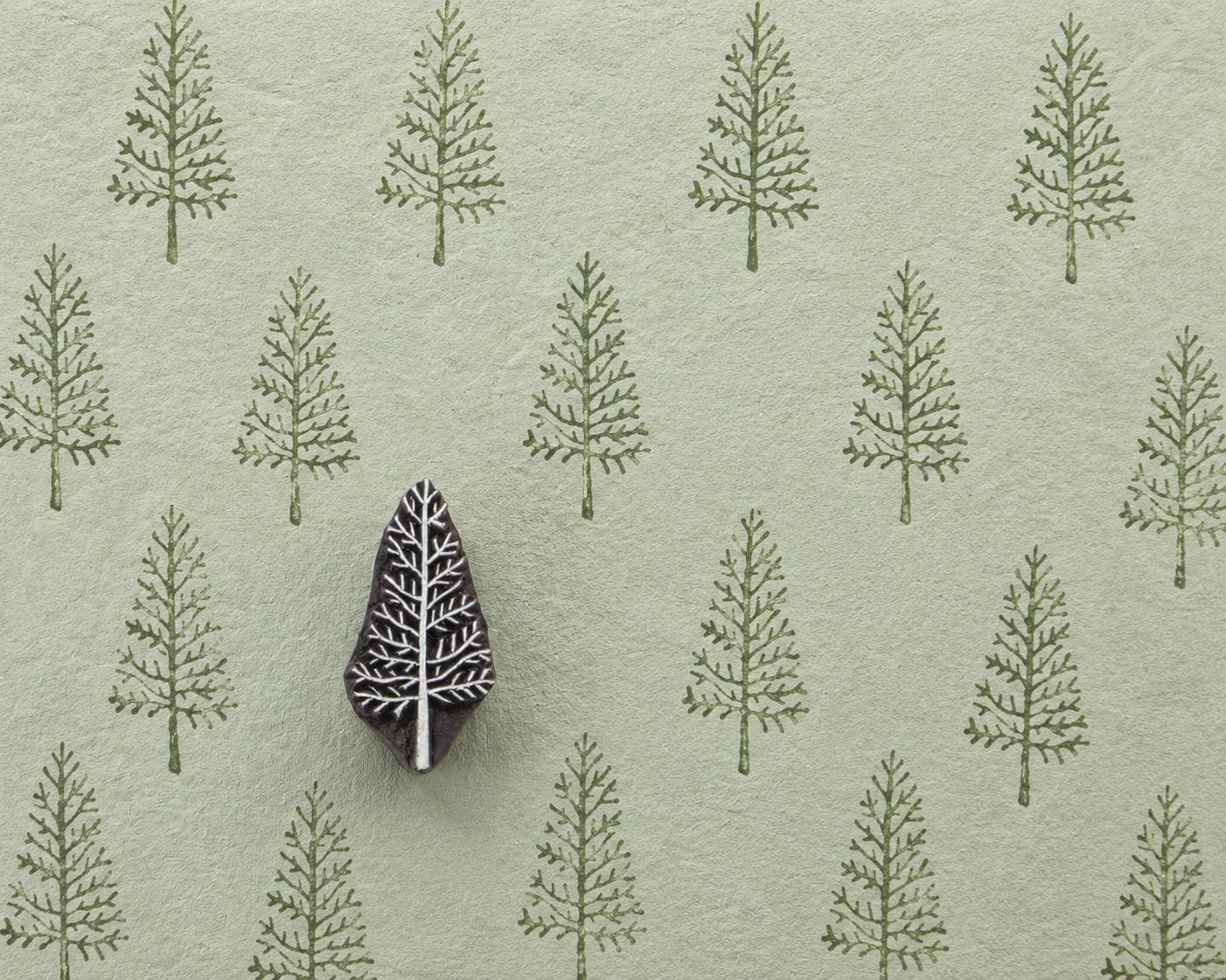 Hand-carved wooden printing block with a slender branching evergreen motif, shown with repeated green impressions on textured handmade paper.
