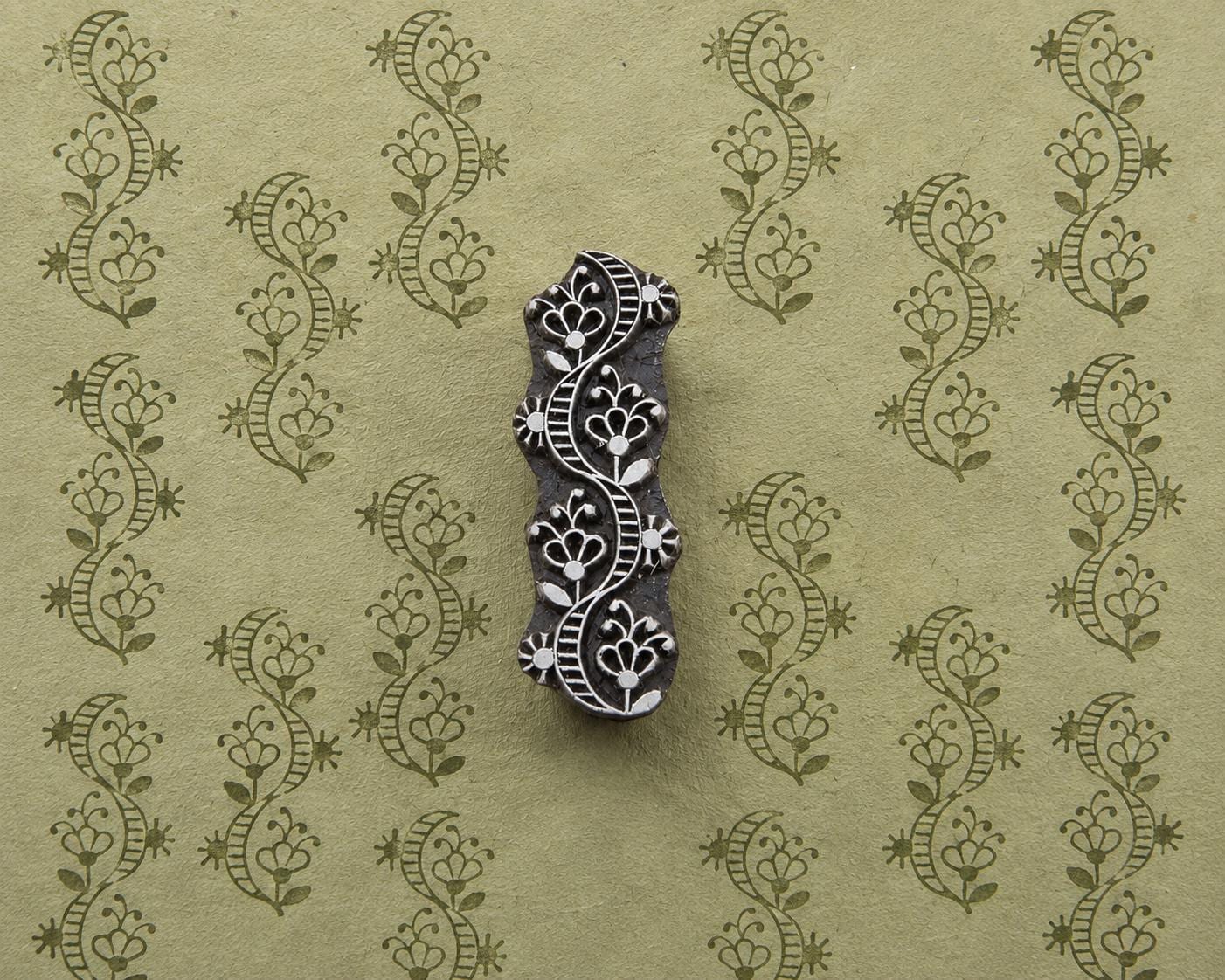 Hand-carved wooden printing block with a flowing floral ornament, shown with repeated green impressions on textured handmade paper.
