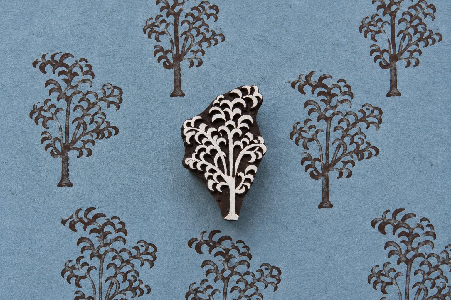 Hand-carved wooden printing block of a tree with a cascading branch and curved foliage motif, shown with repeated brown impressions on blue handmade paper.