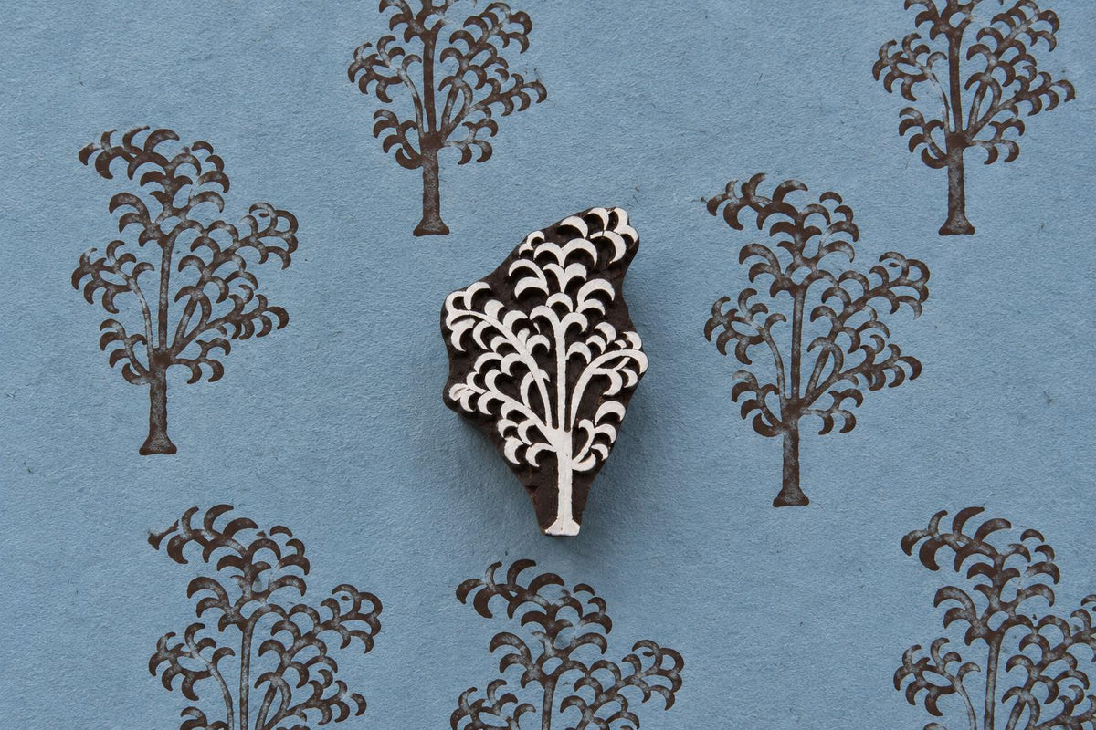 Hand-carved wooden printing block of a tree with a cascading branch and curved foliage motif, shown with repeated brown impressions on blue handmade paper.