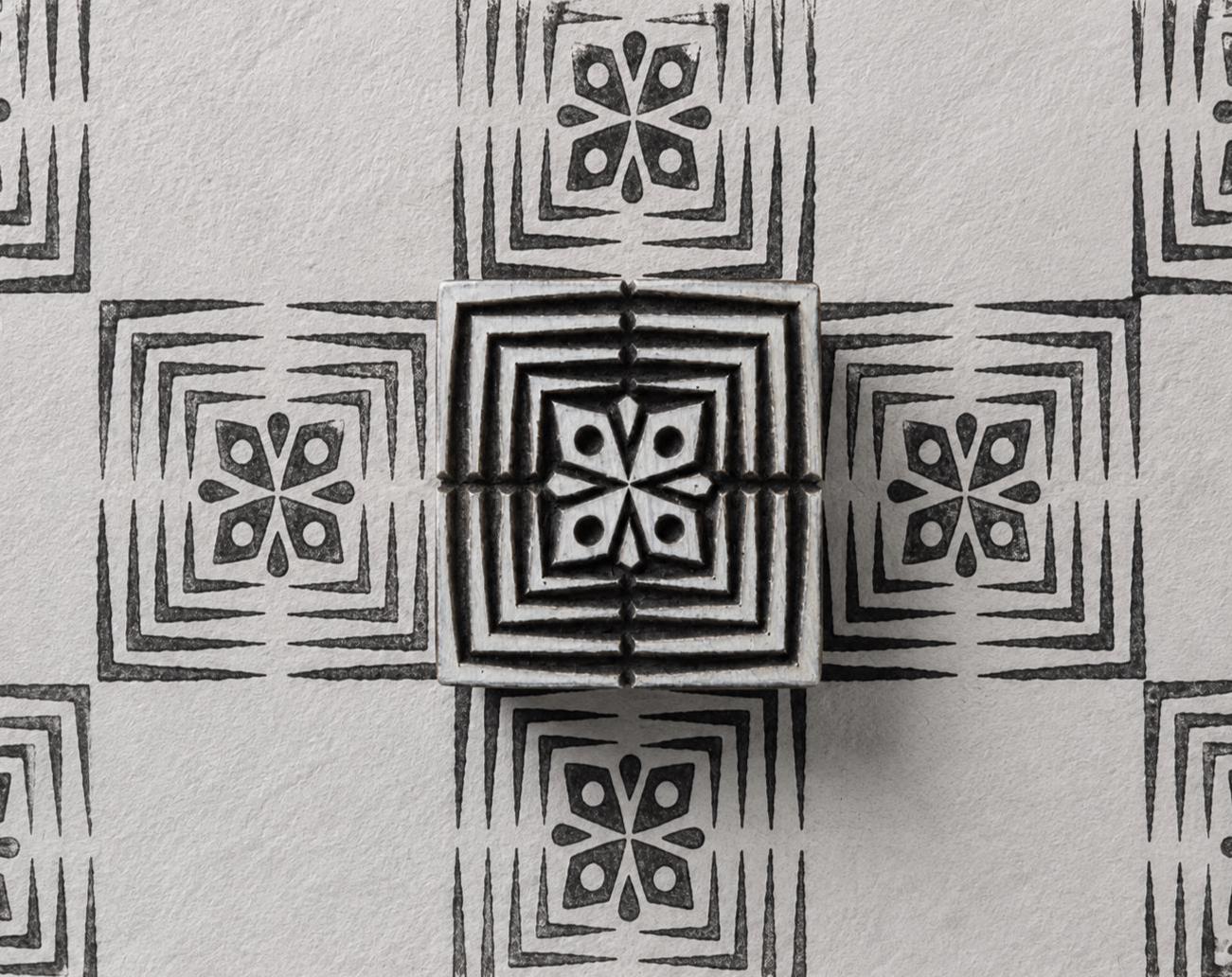 Hand carved Indian wood printing block with four-petal geometric flower and nested square border, shown with a repeating black print on handmade paper