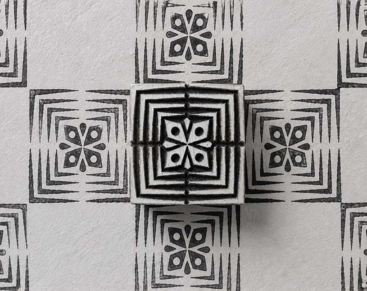 Hand carved Indian wood printing block with four-petal geometric flower and nested square border, shown with a repeating black print on handmade paper