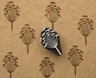 Hand-carved wooden printing block featuring a botanical seed-head motif, shown with repeated brown impressions on textured handmade paper.