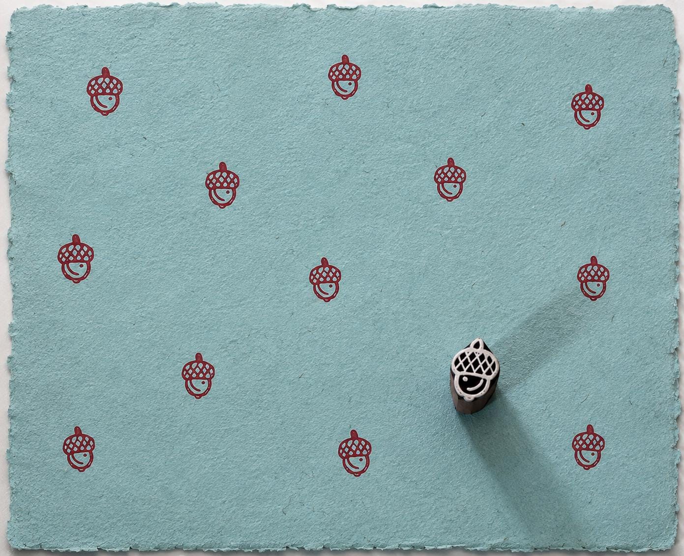 Hand carved acorn wooden printing block creating a repeating red pattern on handmade blue paper