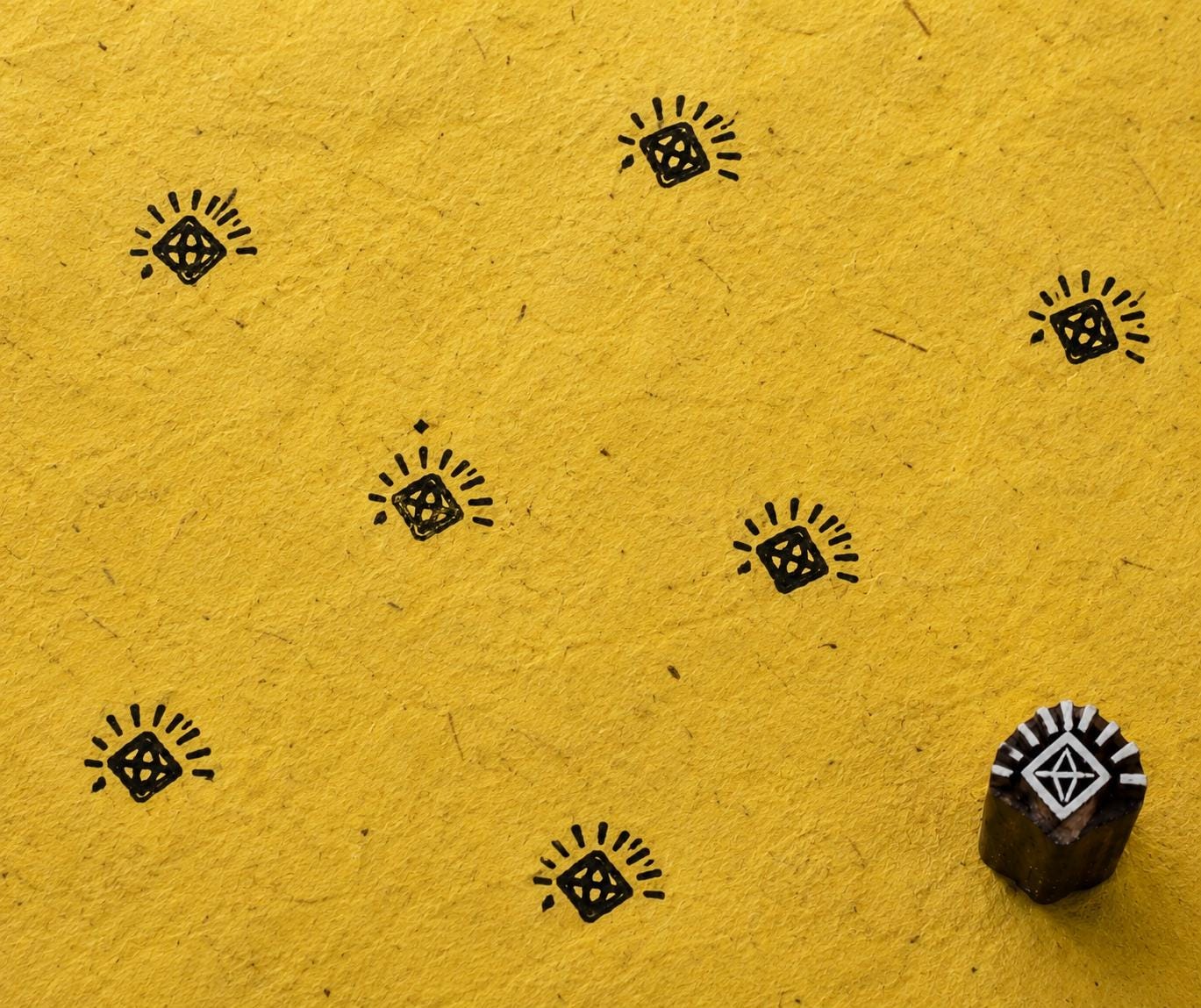 Hand carved geometric diamond wooden printing block with radiating lines shown on textured mustard handmade paper with repeated black impressions.