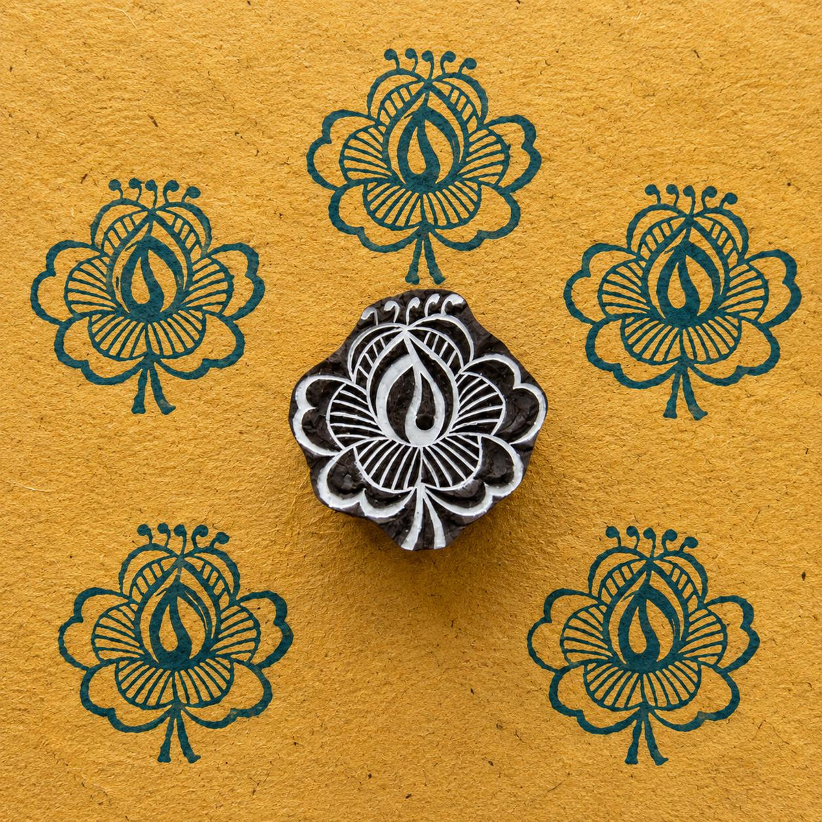 Hand carved wooden Flame Lotus printing block displayed on handmade ochre paper with repeated deep blue floral impressions.