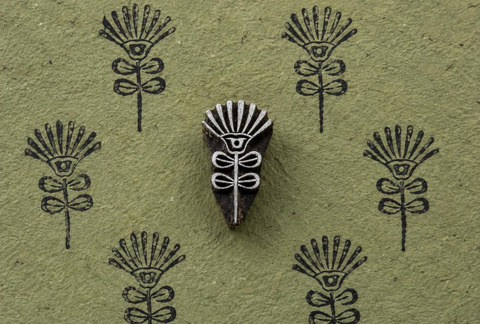 Hand-carved wooden block with a stylized sunburst flower and botanical stem, shown beside repeated impressions on textured green paper.