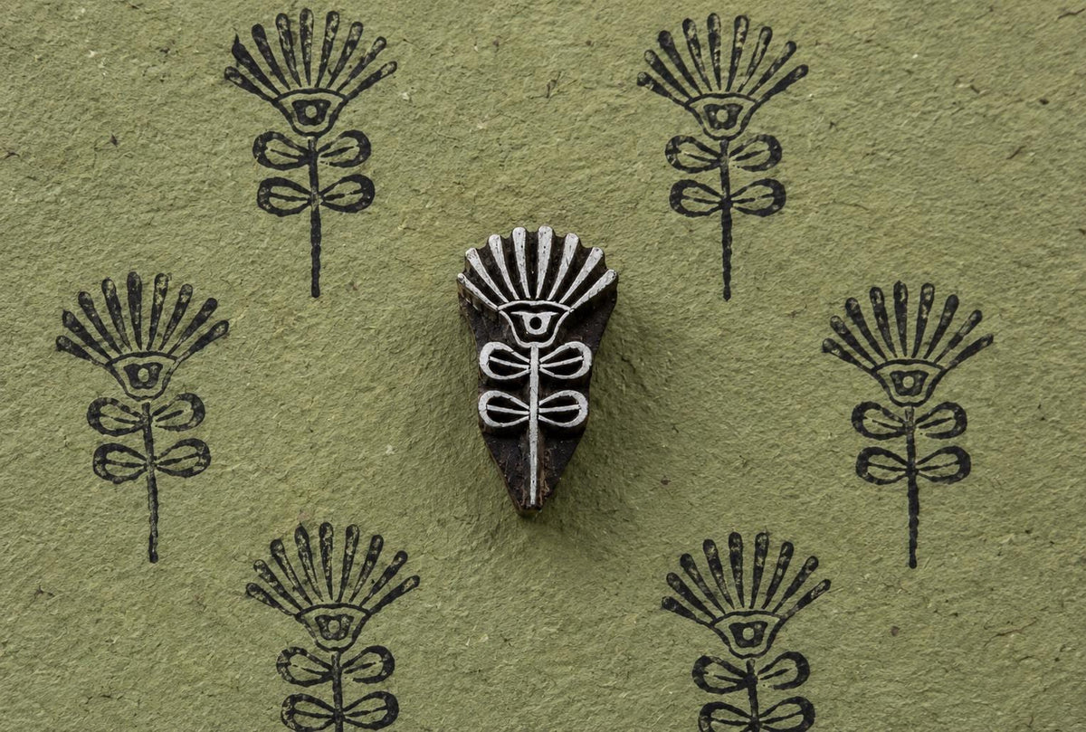 Hand-carved wooden block with a stylized sunburst flower and botanical stem, shown beside repeated impressions on textured green paper.