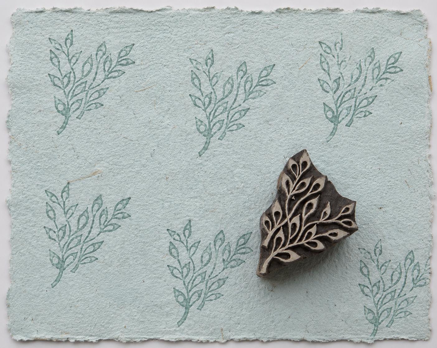 Hand carved leaf sprig wood printing block with repeated botanical impressions on pale blue handmade paper