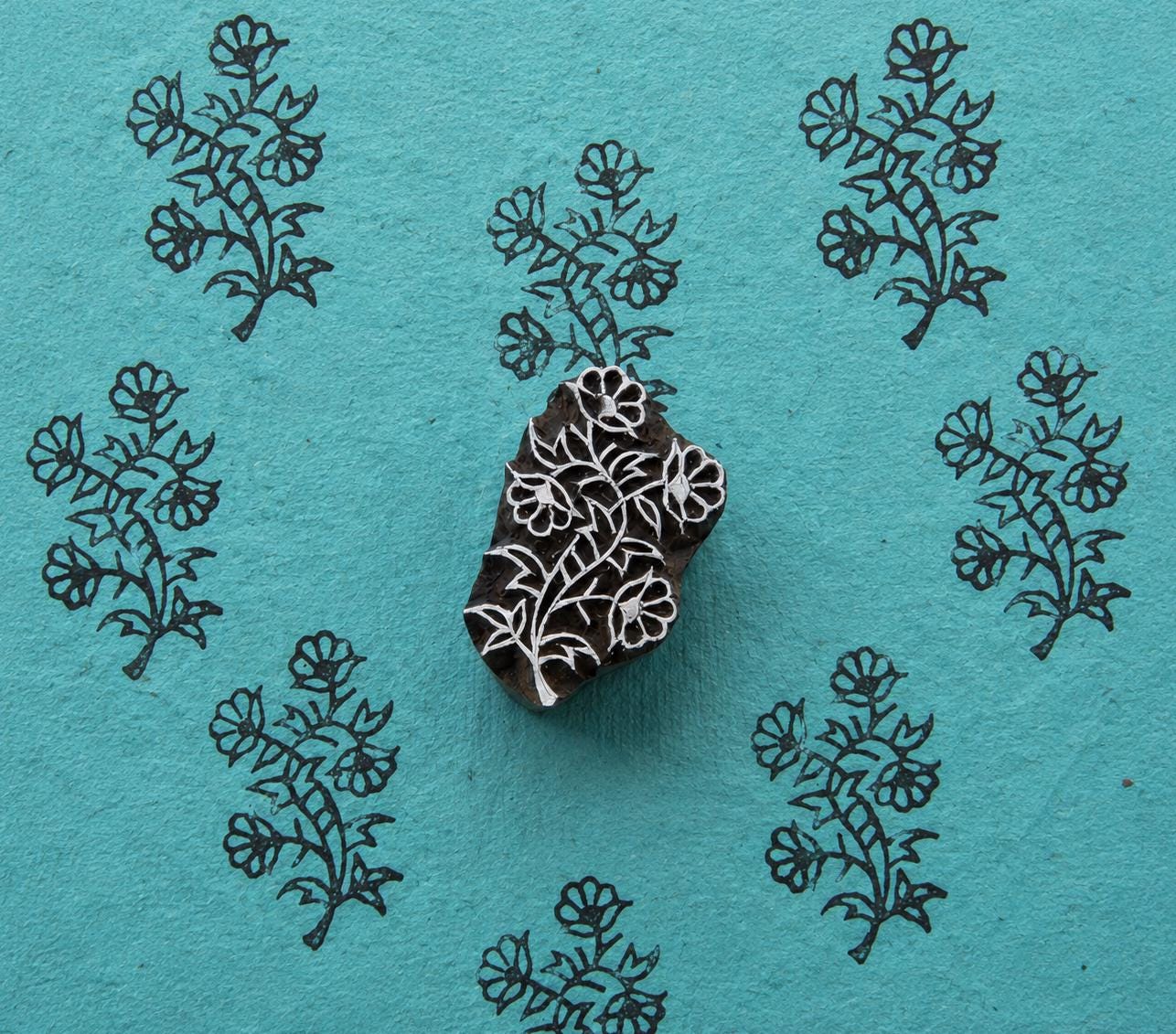 Hand-carved wooden block featuring a leafy wildflower branch, surrounded by repeated floral impressions on textured turquoise handmade paper.