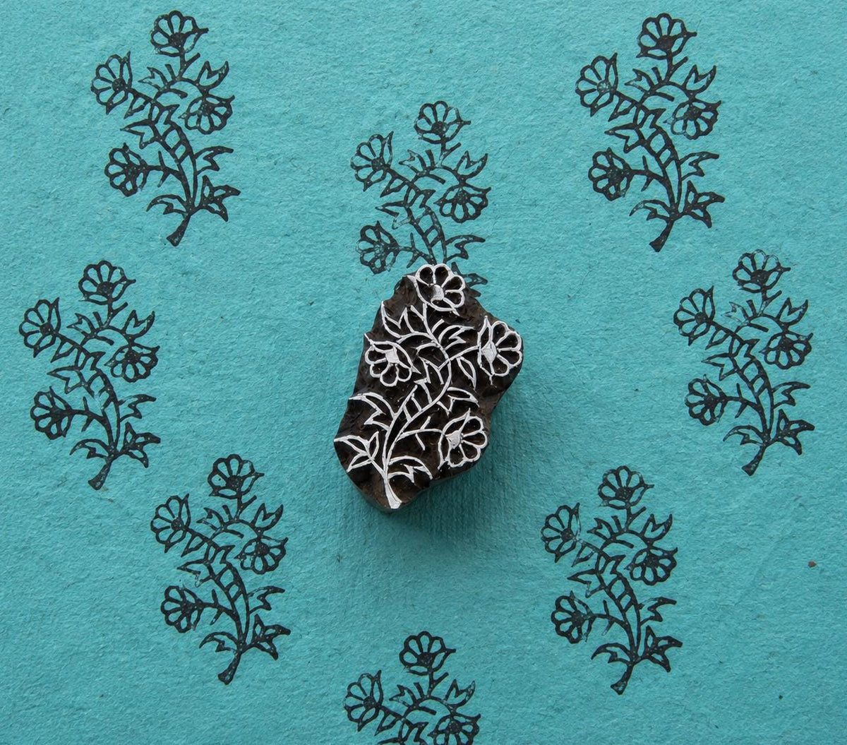 Hand-carved wooden block featuring a leafy wildflower branch, surrounded by repeated floral impressions on textured turquoise handmade paper.