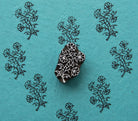 Hand-carved wooden block featuring a leafy wildflower branch, surrounded by repeated floral impressions on textured turquoise handmade paper.