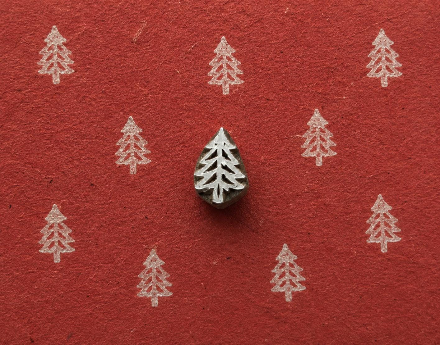 Simple Evergreen wooden stamp shown with repeated white tree impressions on textured red handmade paper.