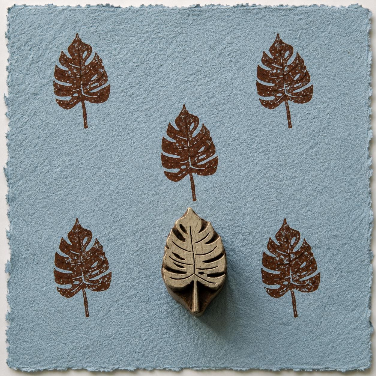Hand carved Monstera split leaf wooden printing block with repeated brown impressions on blue handmade paper