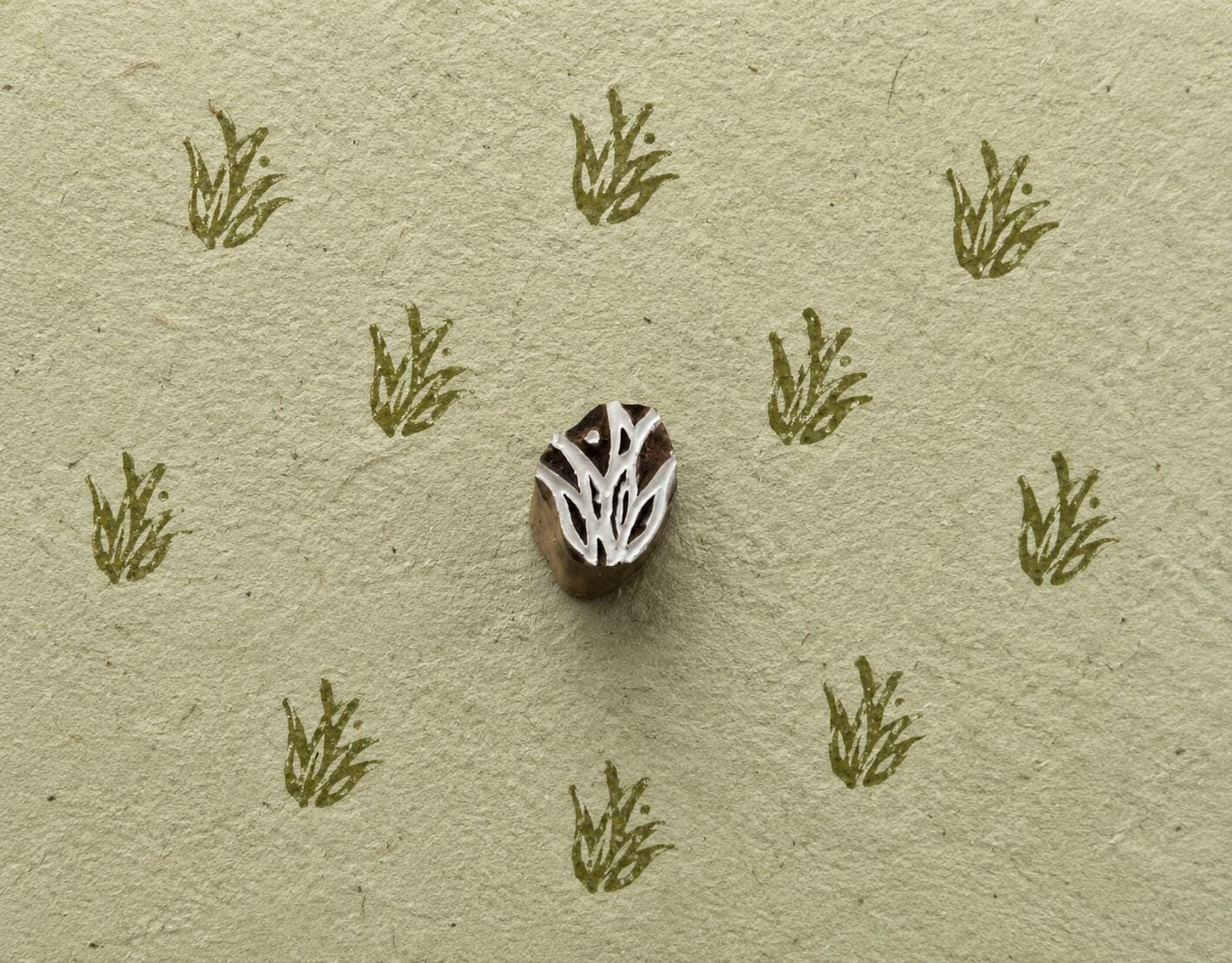 A delicate botanical motif inspired by a small tuft of wild grass, hand carved into wood with fine, organic lines and tiny dot details.