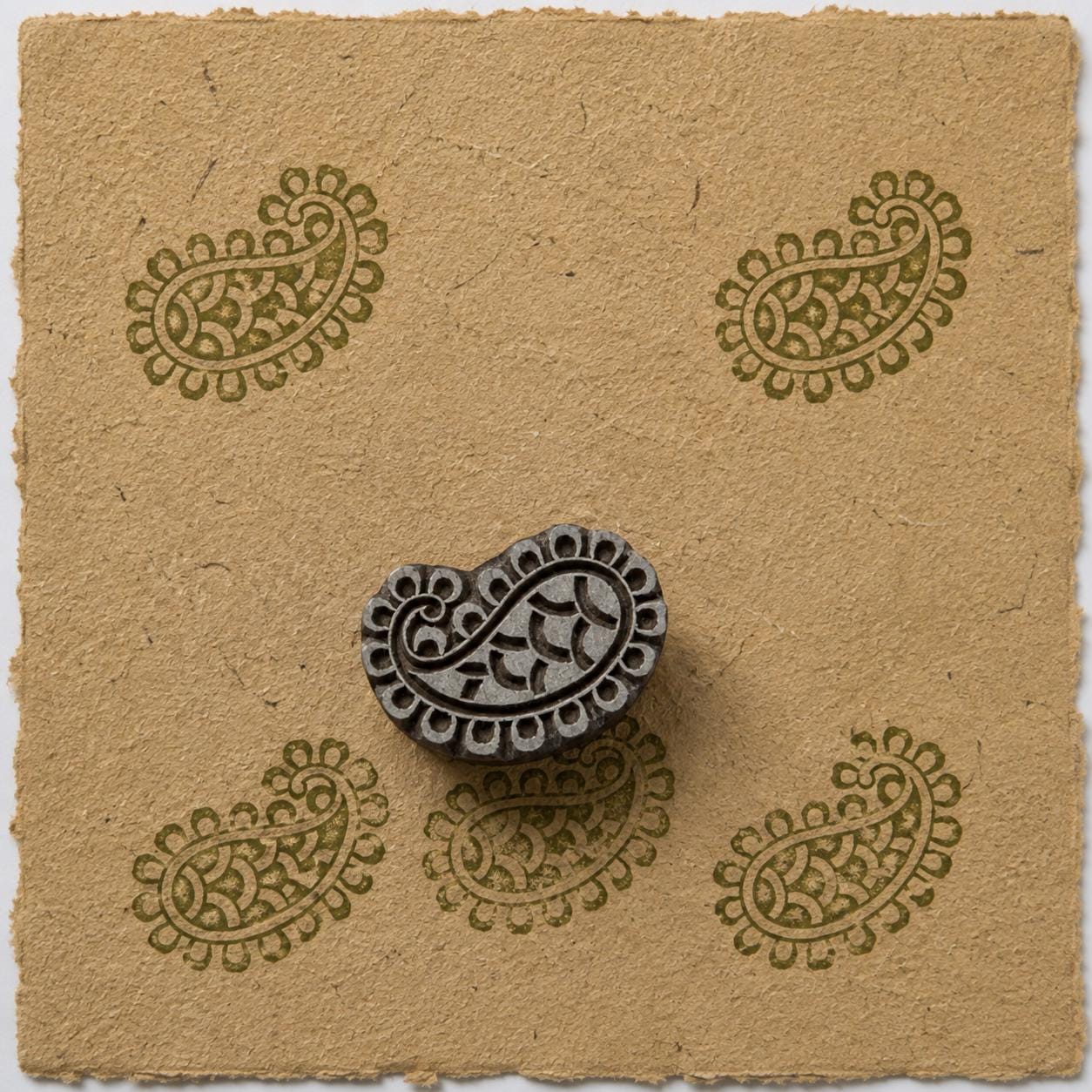 Hand carved Indian wooden printing block featuring a traditional heritage paisley motif with decorative inner details and a rounded border, shown with repeated green impressions on handmade paper.