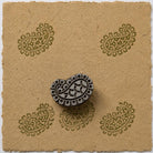 Hand carved Indian wooden printing block featuring a traditional heritage paisley motif with decorative inner details and a rounded border, shown with repeated green impressions on handmade paper.