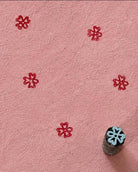 Hand carved Indian wooden printing block with a small petal floral motif, shown creating a repeating red pattern on handmade pink paper.