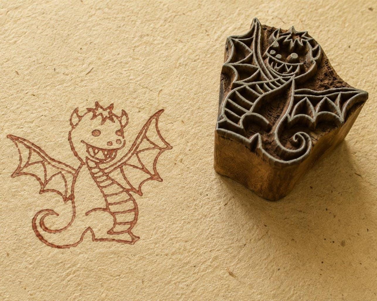 Hand carved whimsical baby dragon wooden printing block shown beside its printed dragon impression on handmade textured paper