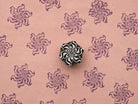 Hand carved curled petal flower wood stamp surrounded by repeated purple floral impressions on textured pink handmade paper.