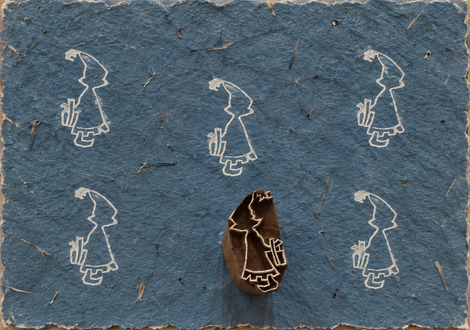 Hand carved Indian wooden printing block showing a little girl holding a gift behind her back, surrounded by printed impressions on blue handmade paper.