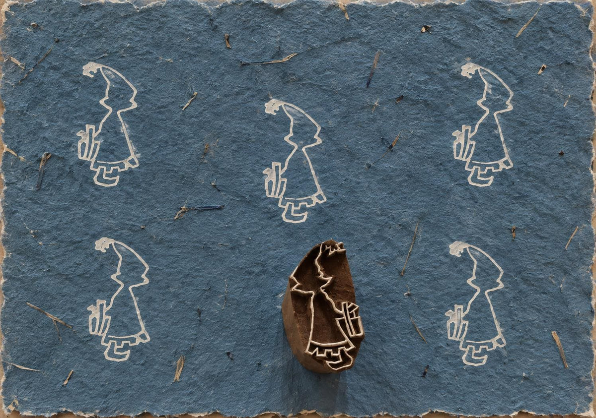 Hand carved Indian wooden printing block showing a little girl holding a gift behind her back, surrounded by printed impressions on blue handmade paper.