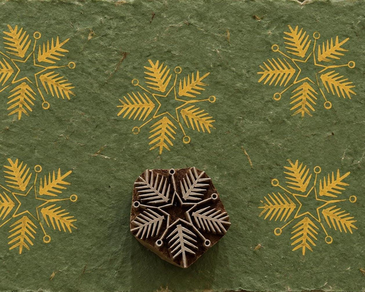 Hand carved wooden botanical snowflake printing block surrounded by repeated mustard yellow snowflake prints on textured green handmade paper.