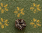Hand carved wooden botanical snowflake printing block surrounded by repeated mustard yellow snowflake prints on textured green handmade paper.