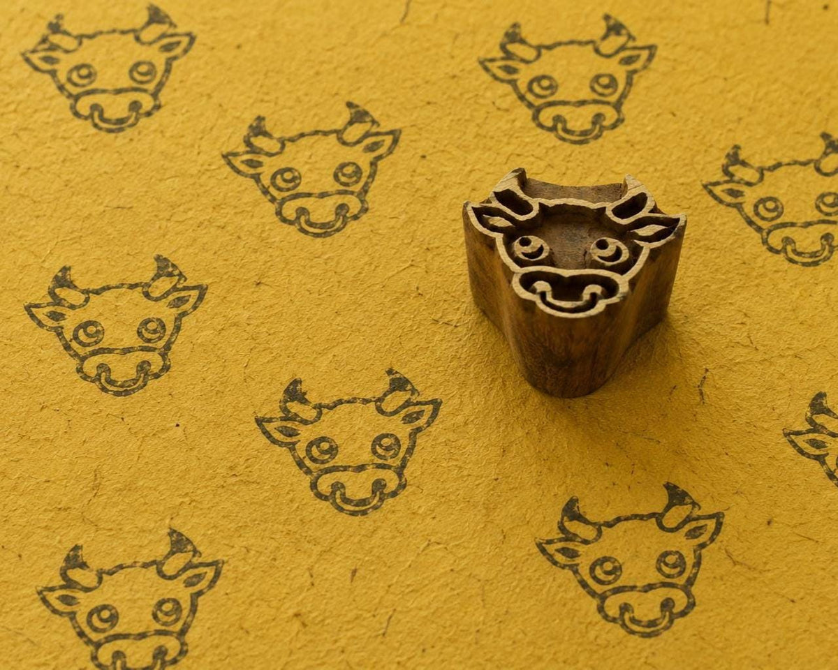 Hand carved wooden baby cow face printing block surrounded by repeated cow face prints on textured golden yellow handmade paper.