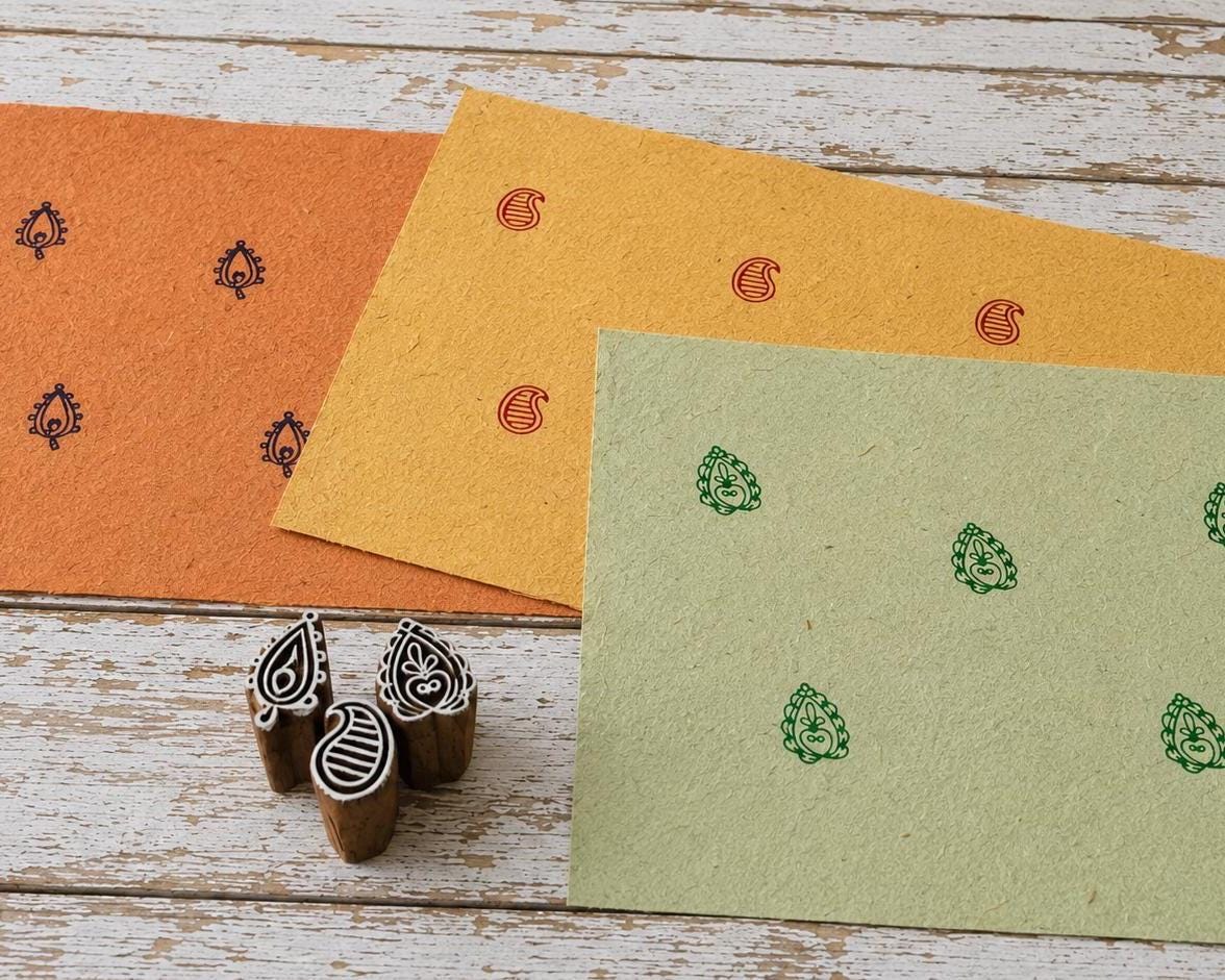 Set of three small hand carved Indian wooden printing blocks with three different printed motifs displayed on textured orange, yellow and green handmade paper.