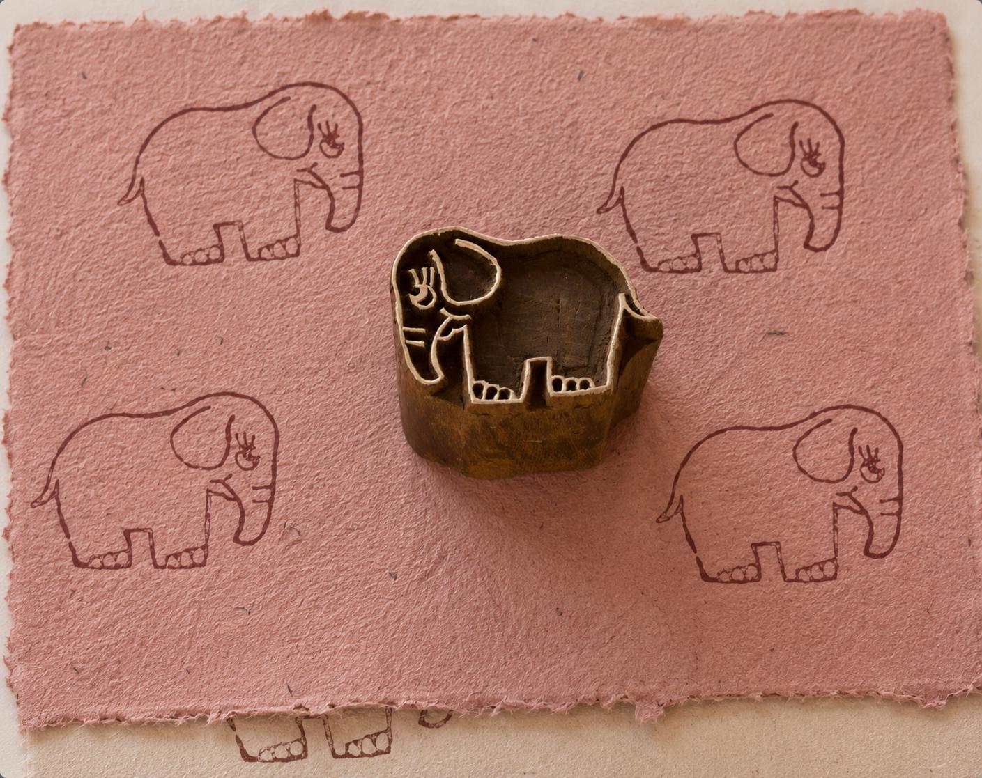 Hand carved Happy Elephant wooden printing block surrounded by repeated elephant impressions on textured dusty pink handmade paper