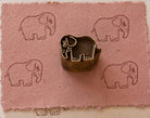 Hand carved Happy Elephant wooden printing block surrounded by repeated elephant impressions on textured dusty pink handmade paper