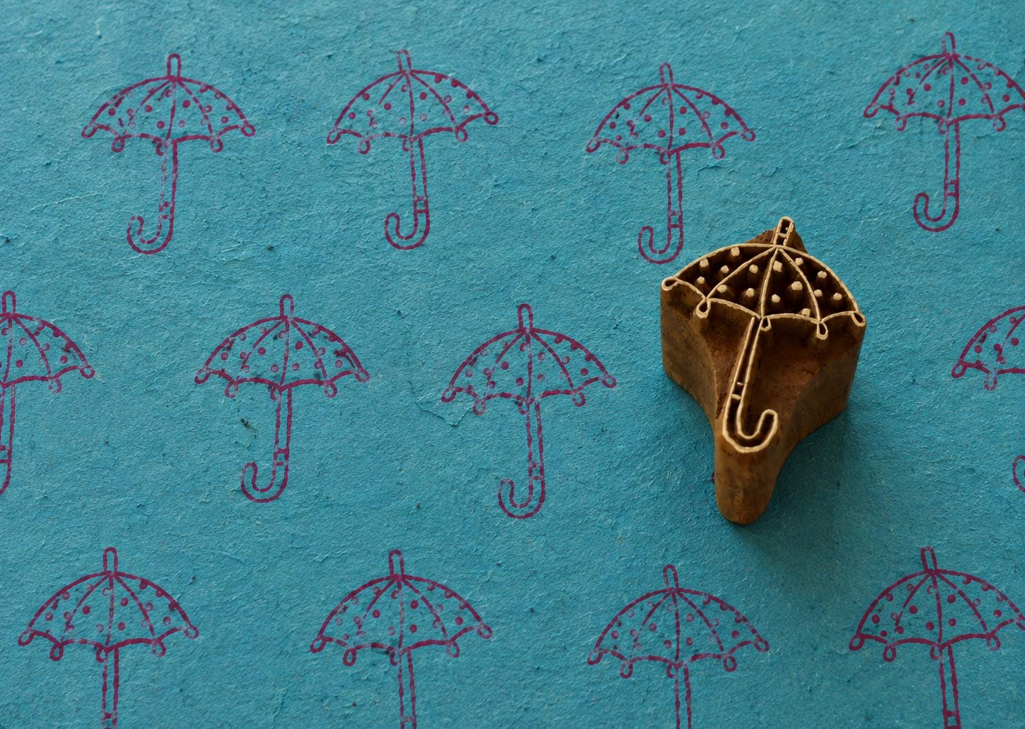 Hand carved Raindrop Umbrella wooden printing block with repeated umbrella impressions on textured teal handmade paper