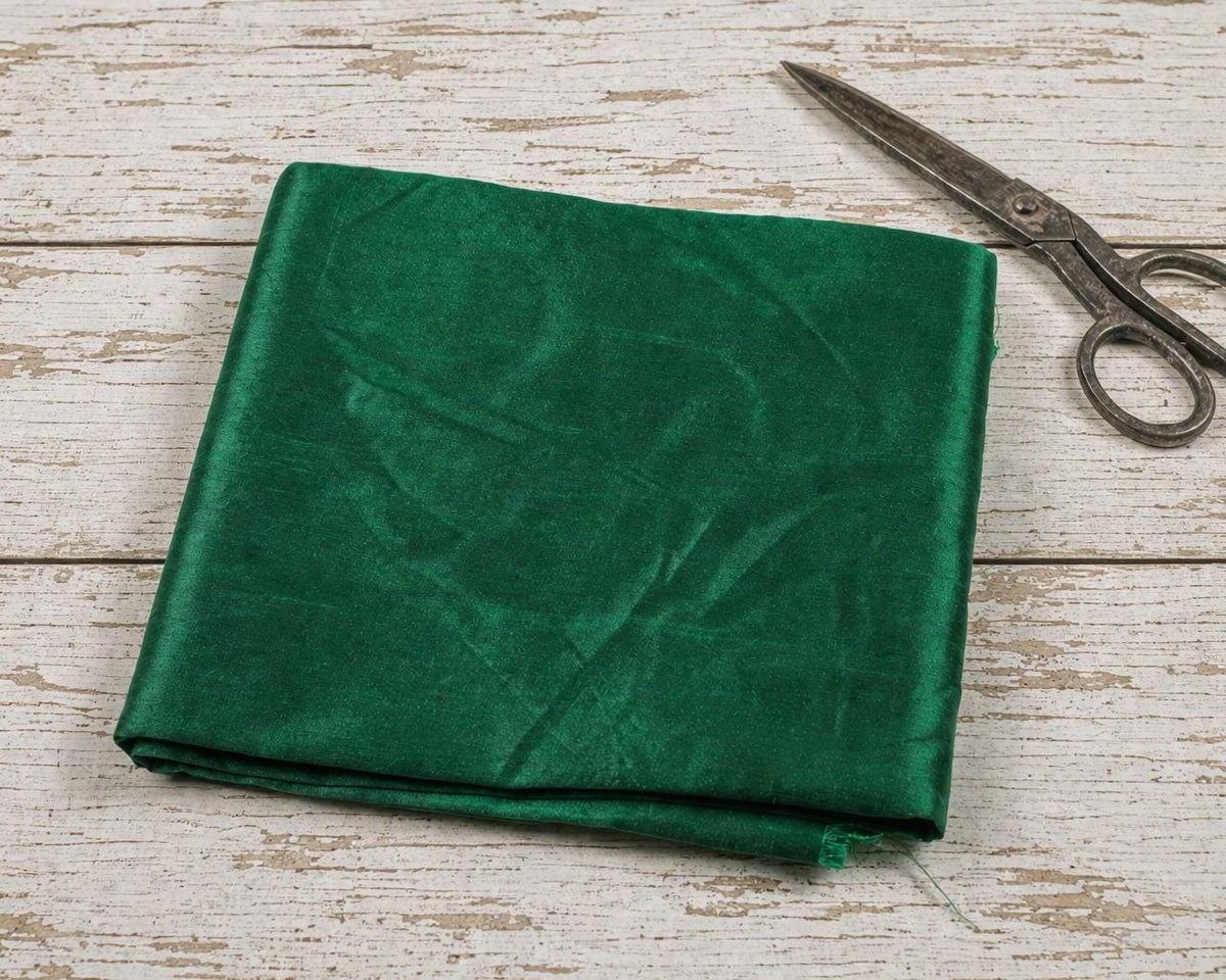 Emerald green faux silk dupioni fabric folded on a distressed white wooden table beside vintage metal scissors.
