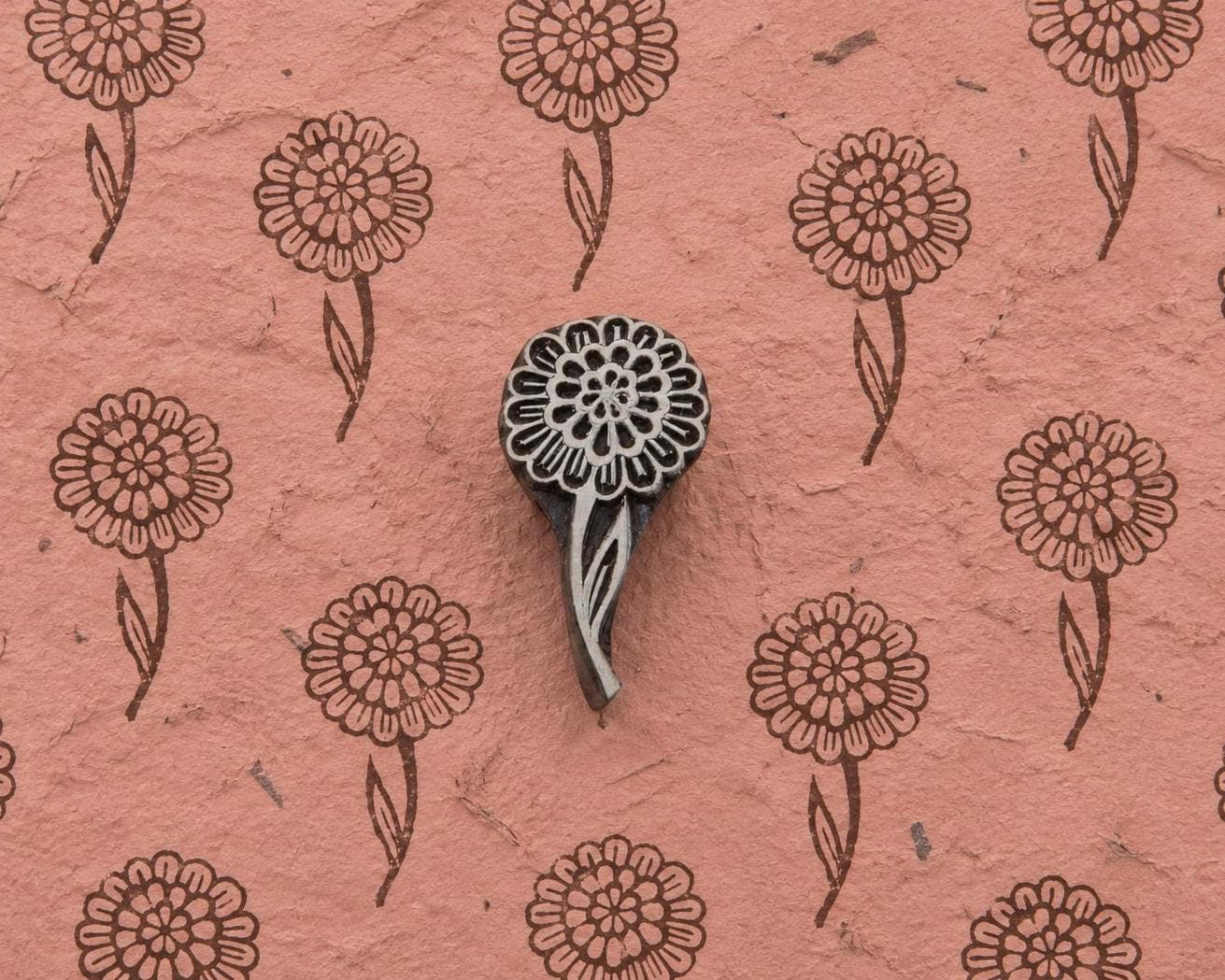 Layered Bloom hand carved wooden printing block with repeated round floral impressions on textured handmade pink paper.