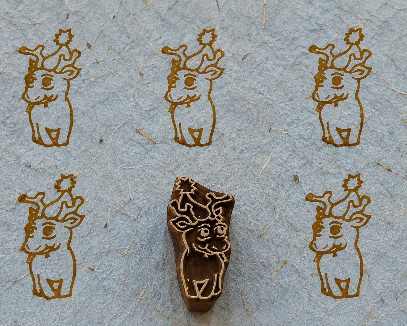Hand carved wooden baby reindeer printing block surrounded by five brown reindeer prints on textured grey handmade paper.