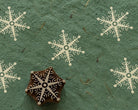 Hand carved wooden printing block featuring a decorative dotwork snowflake, surrounded by repeated cream snowflake impressions on textured green handmade paper.
