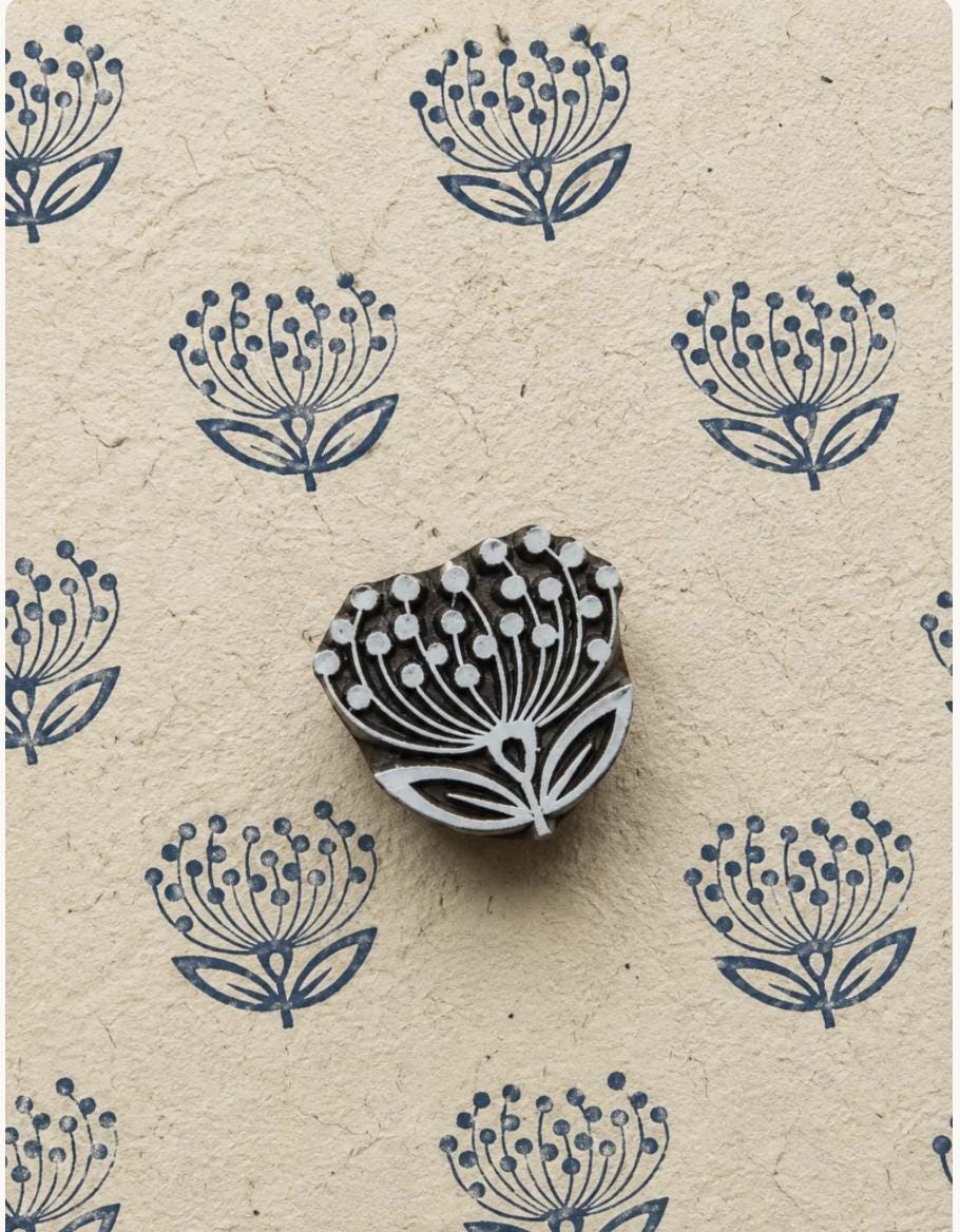 Hand carved wooden dandelion-inspired motif shown with repeated blue impressions on textured handmade paper.