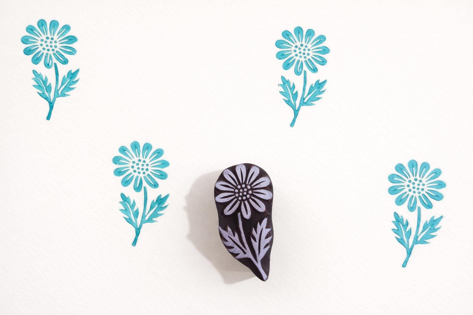 Hand carved wooden daisy botanical block shown with repeated turquoise impressions on white handmade paper.