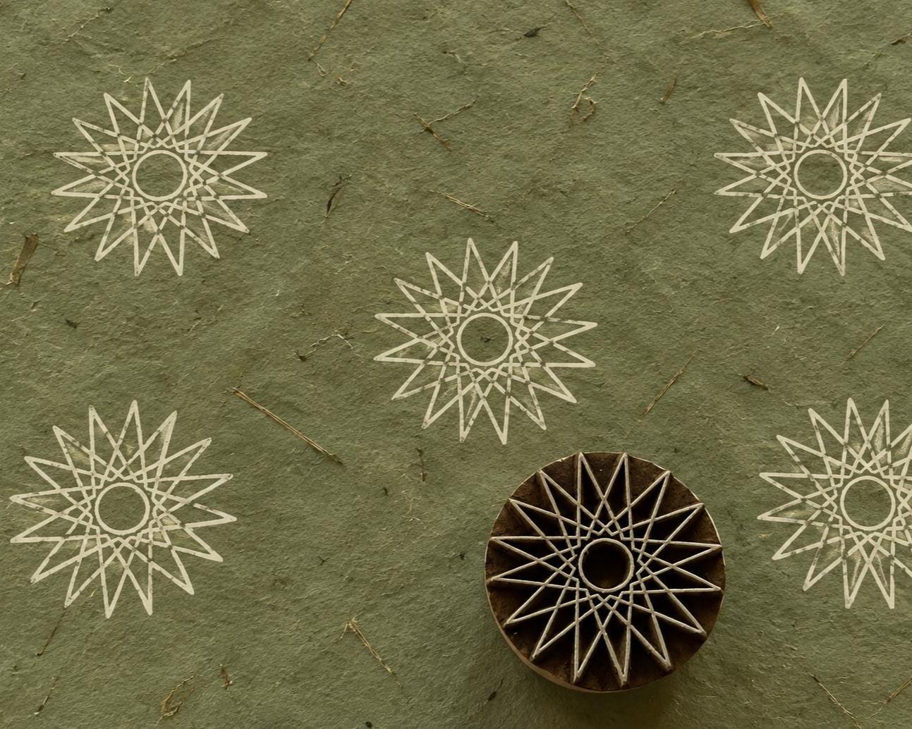 Hand carved Indian wooden printing block with a geometric sunburst and star motif, shown with repeated prints on textured green handmade paper.