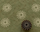 Hand carved Indian wooden printing block with a geometric sunburst and star motif, shown with repeated prints on textured green handmade paper.