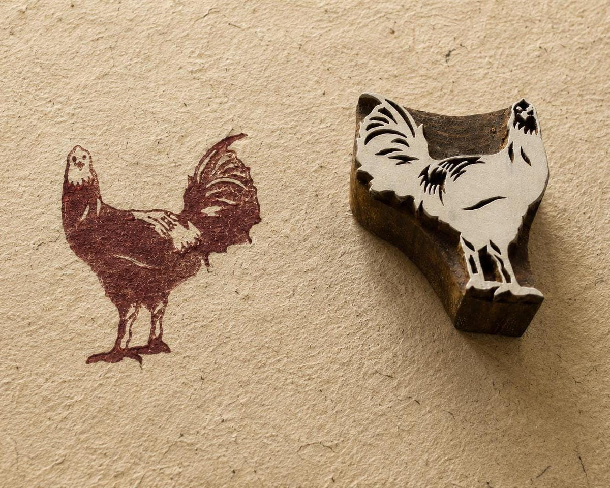 Hand carved Indian wooden printing block with a rooster design shown beside its rustic brown printed impression on textured handmade paper.