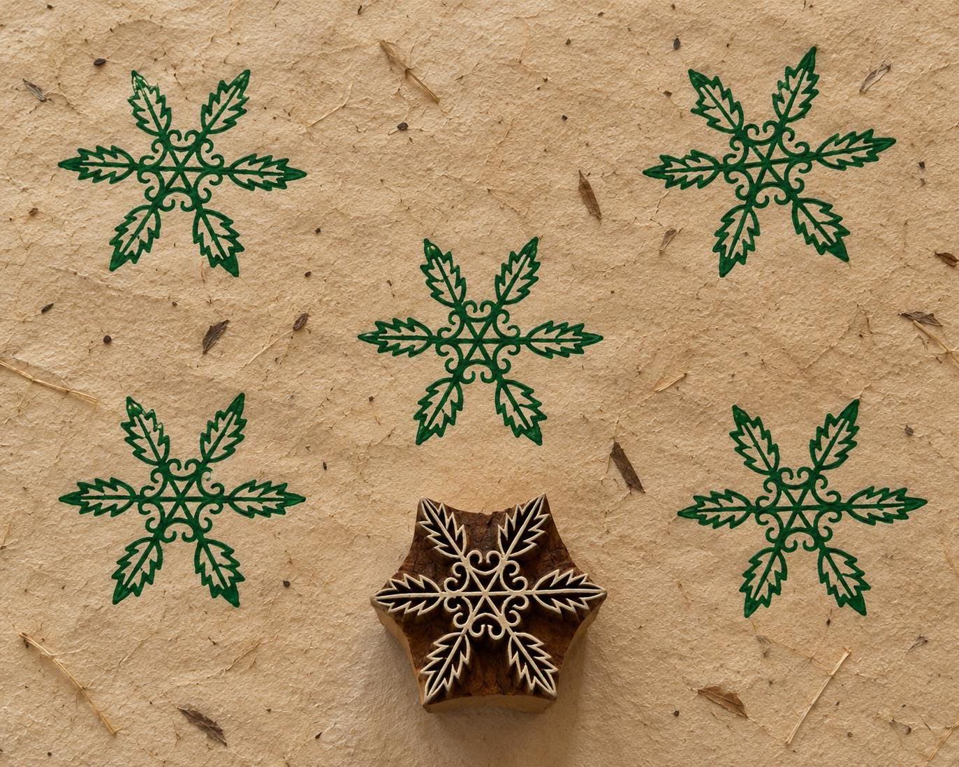 Hand carved wooden printing block featuring a six-leaf botanical snowflake design, with green printed impressions on textured handmade paper.
