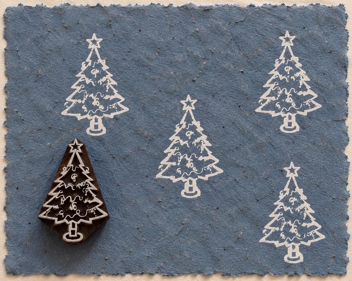 Hand carved wooden printing block featuring a decorated Christmas tree with a star topper, shown with white impressions on blue handmade paper.