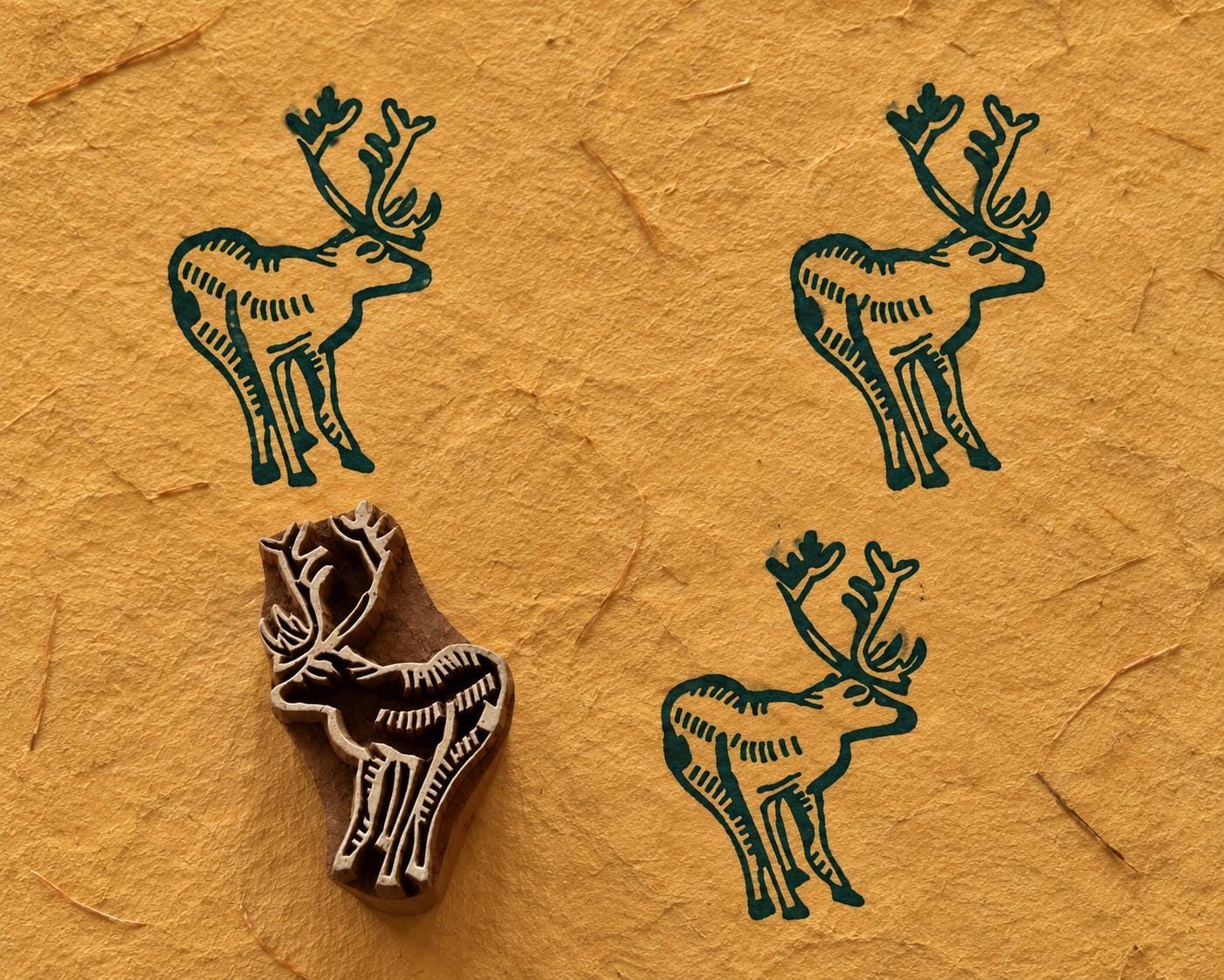 Hand carved wooden printing block of an antlered stag with three dark green printed impressions on mustard handmade paper.
