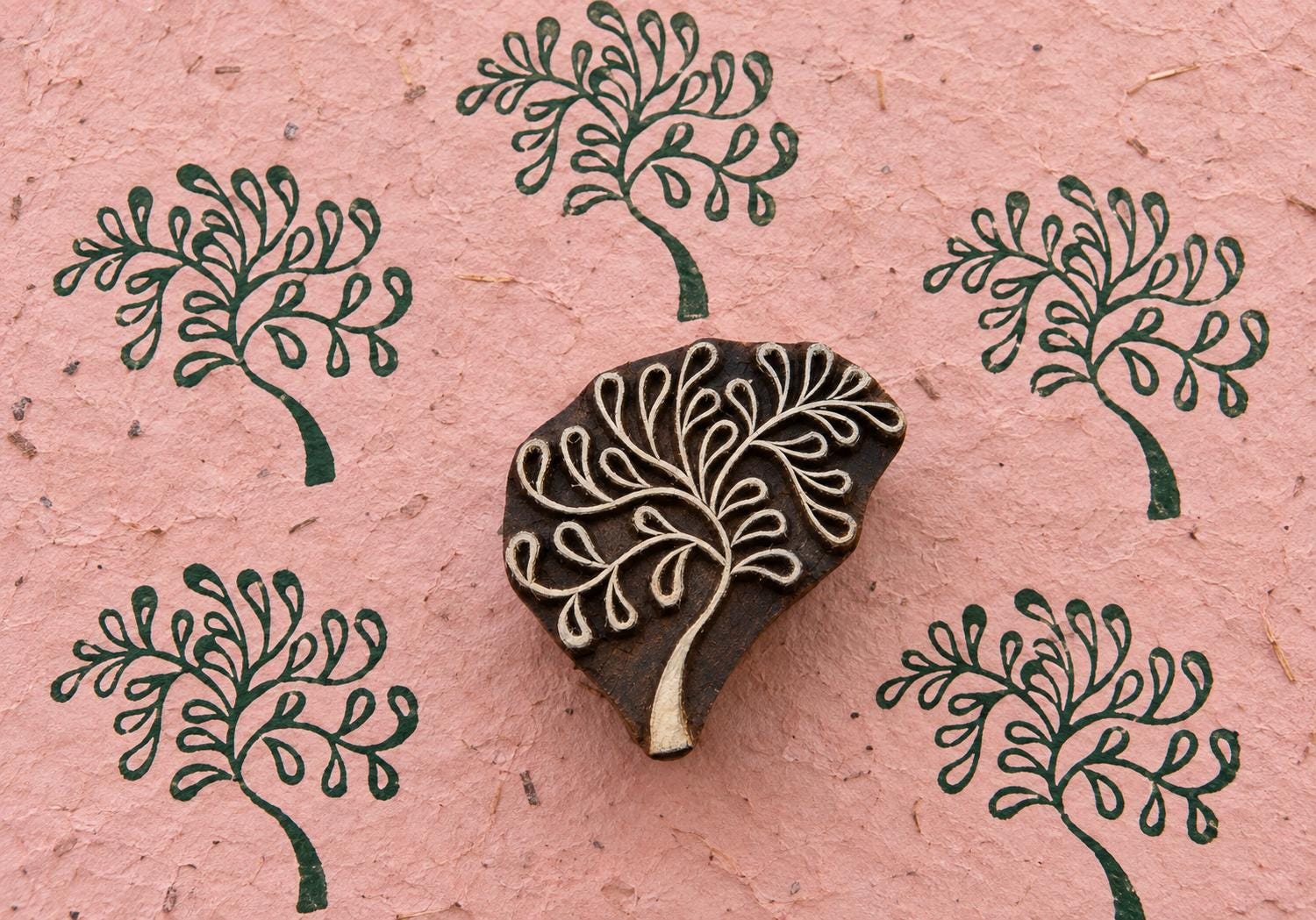 Hand carved wooden Fountain Tree printing block surrounded by repeated dark green tree impressions on textured pink handmade paper.