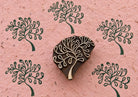 Hand carved wooden Fountain Tree printing block surrounded by repeated dark green tree impressions on textured pink handmade paper.