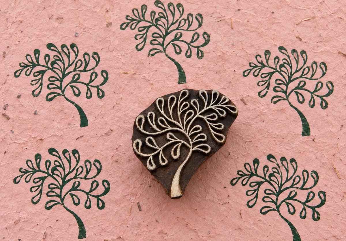 Hand carved wooden Fountain Tree printing block surrounded by repeated dark green tree impressions on textured pink handmade paper.