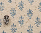 Hand carved Indian wooden printing block with an ornamental heart-shaped motif and geometric interior detail, shown with repeated blue impressions on handmade paper.