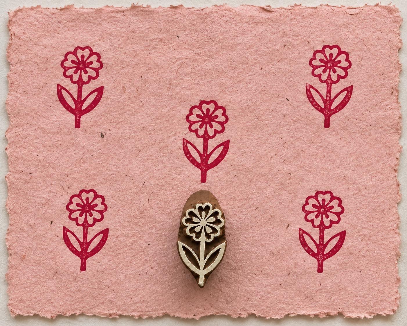 Hand carved wooden Daisy Stem printing block with repeated burgundy floral impressions on pink handmade paper.